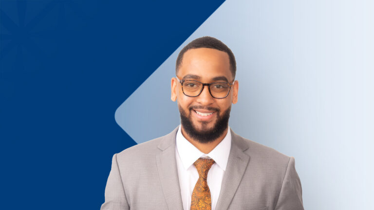 Smiling man in a gray suit and orange patterned tie, wearing glasses, posed against a blue gradient background with geometric shapes.